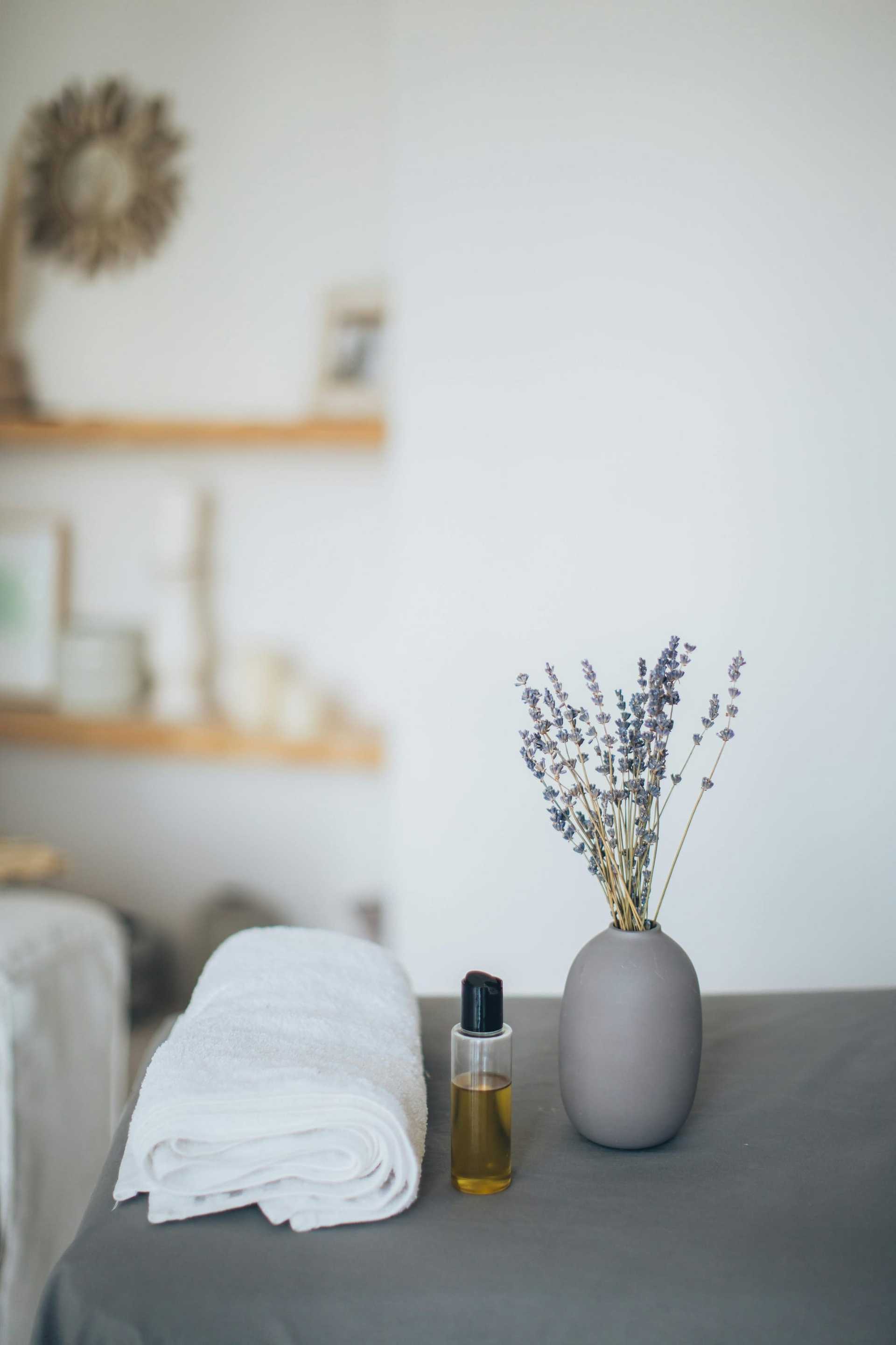 Spa scene with lavender in vase, folded towels, and a bottle of essential oil on a table.