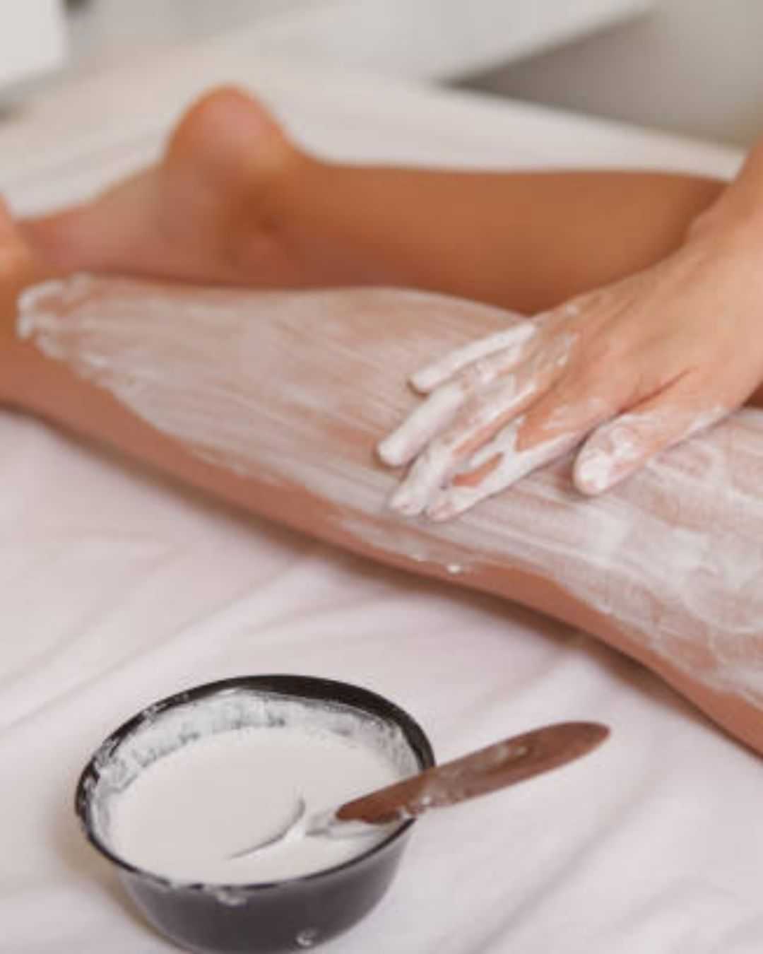 Person applying cream to leg, with a bowl of cream nearby on a white surface.