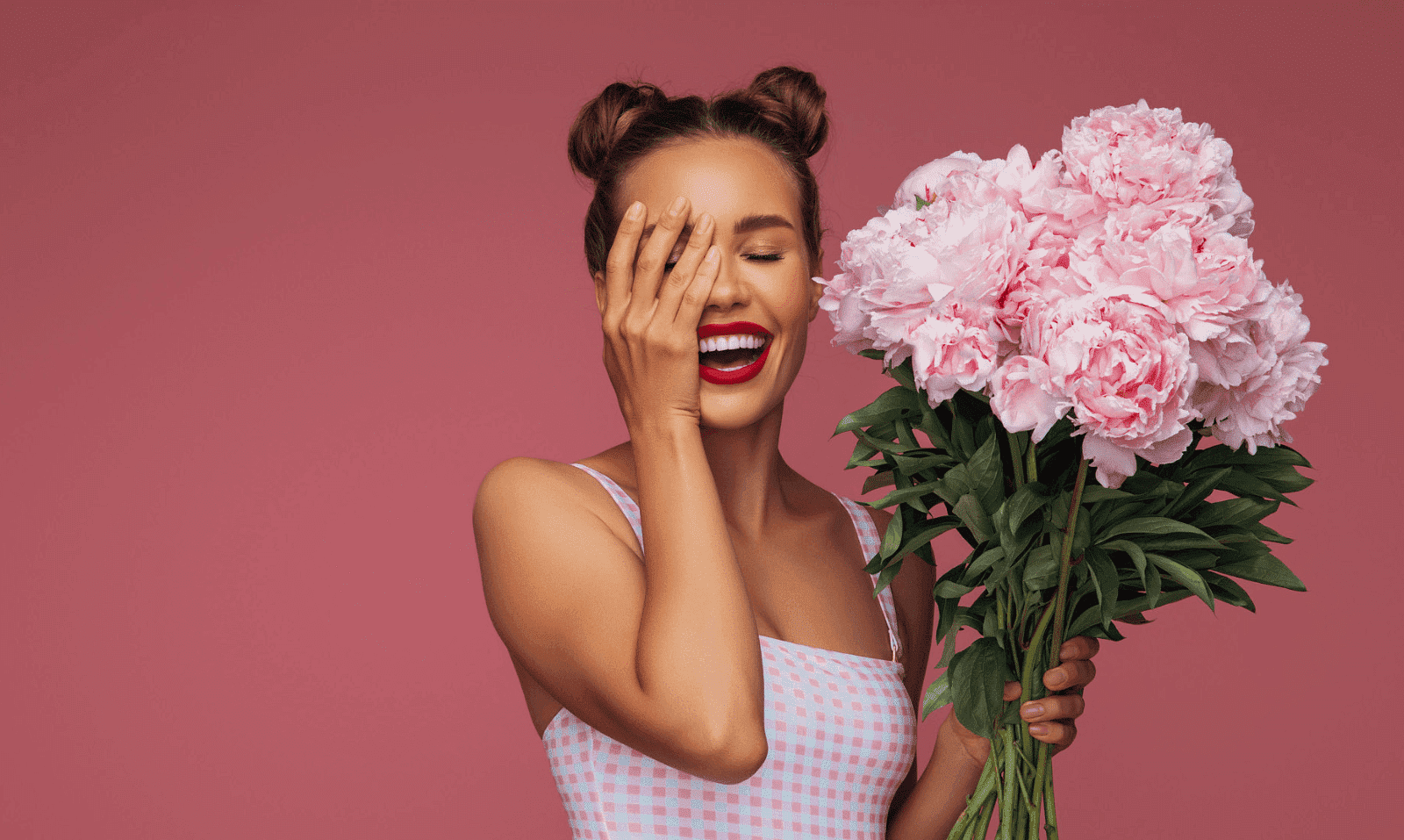Woman laughing, holding pink peonies against pink background.