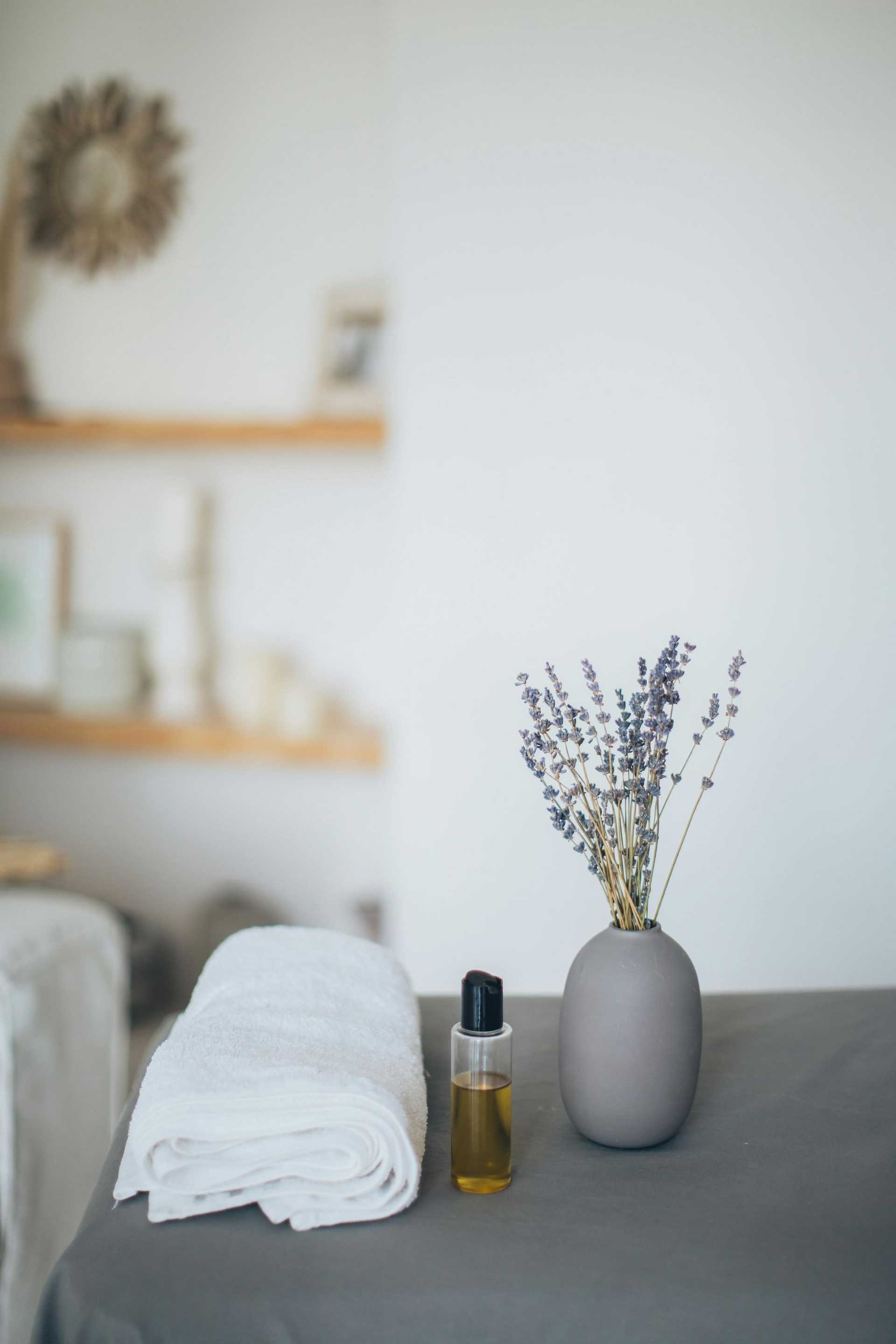 Spa scene with lavender in vase, folded towels, and a bottle of essential oil on a table.