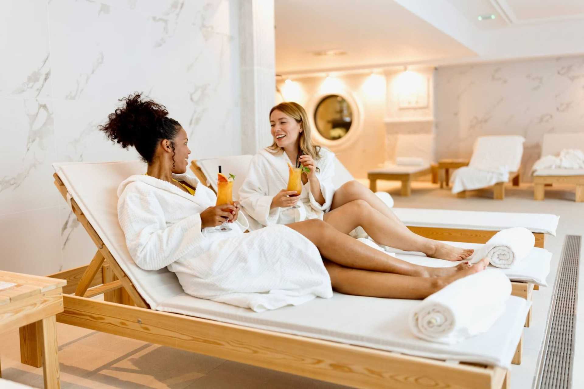 Two women in robes relax on spa loungers, chatting and enjoying drinks.