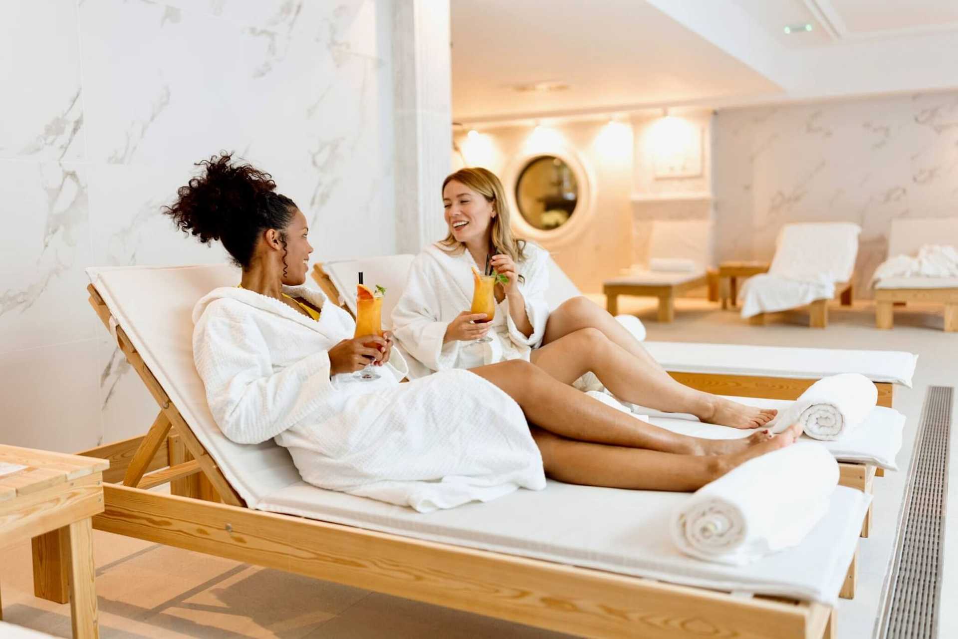 Two women in robes relax on spa loungers, chatting and enjoying drinks.