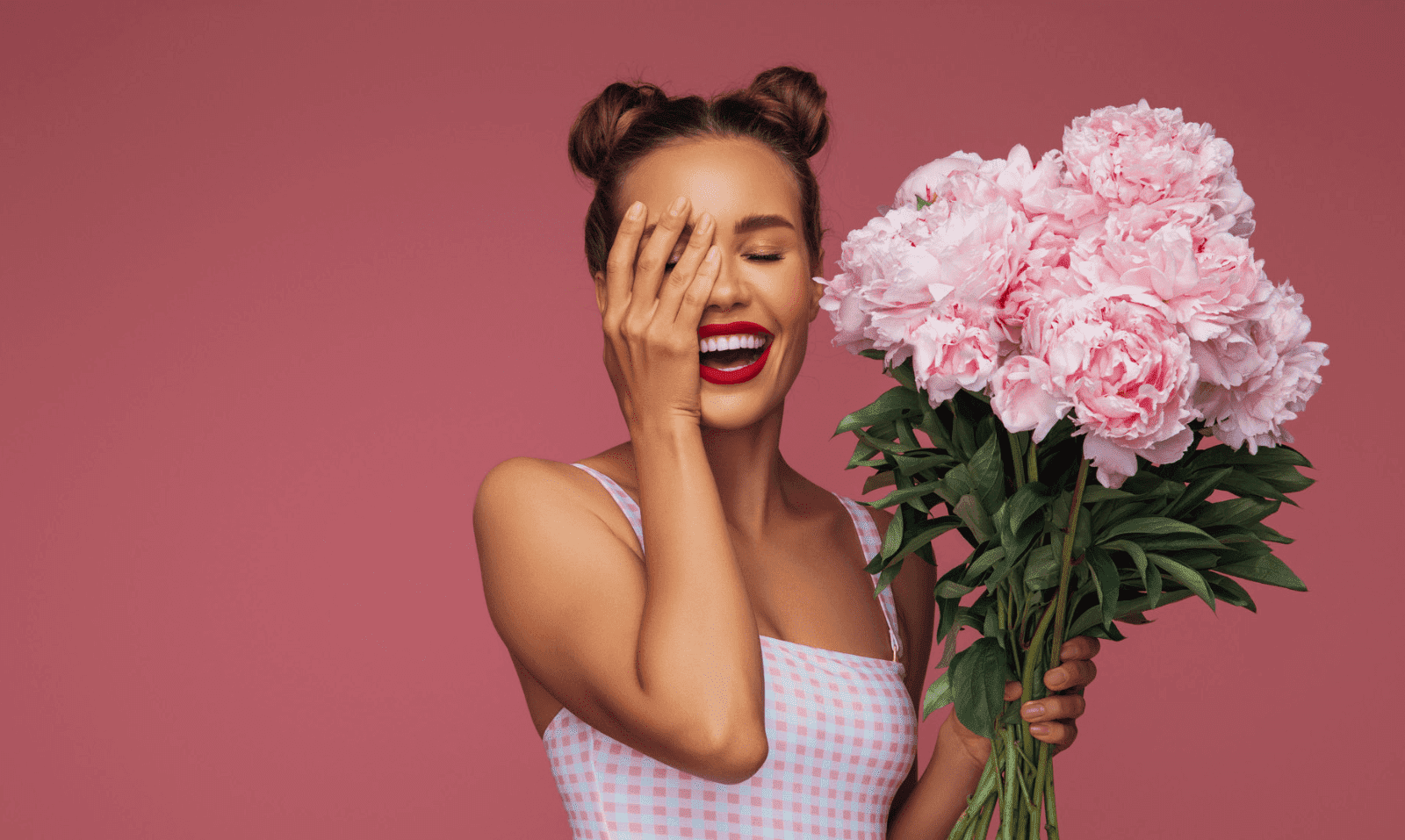 Woman laughing, holding pink peonies against pink background.