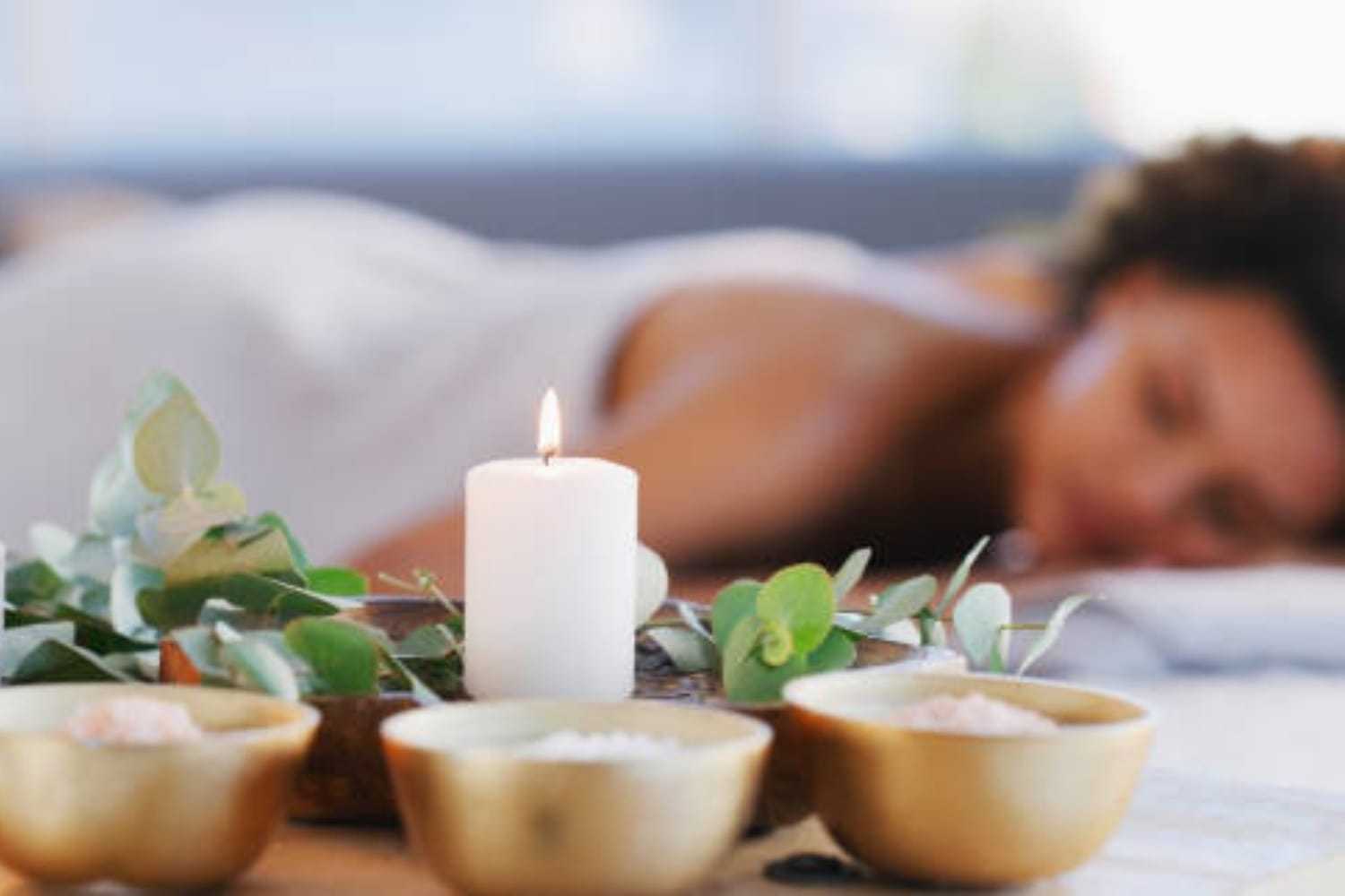 Relaxing spa scene with lit candle, eucalyptus, and person lying down in the background.