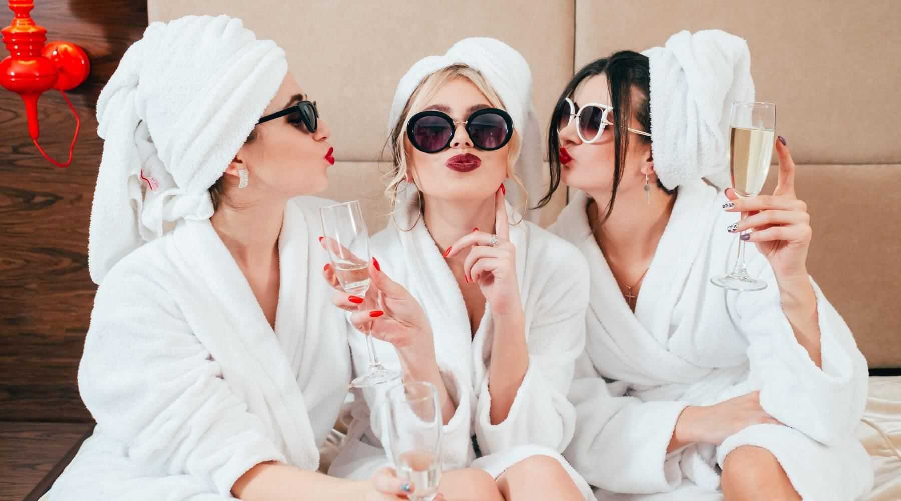 Three women in robes and sunglasses, sipping champagne and posing playfully.